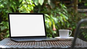tela em branco do laptop maquete e caneca de café branca na mesa de ferro no fundo da árvore do jardim, verde. foto