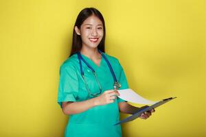 retrato do uma lindo jovem mulher dentro uma amarelo fundo, ásia mulher vestindo uma médico uniforme faz uma sorridente expressão, uma jovem mulher vestindo uma médico estetoscópio tem uma amigáveis expressão. foto