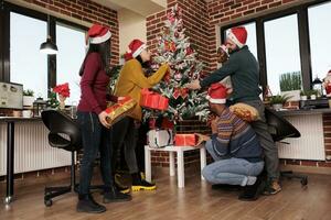 diverso colegas partilha natal apresenta às inverno sazonal feriado festa dentro decorado escritório. feliz colegas de trabalho dentro santa chapéus trocando presentes perto Natal árvore com enfeites foto