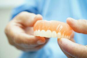 ásia Senior mulher paciente segurando dentes dentadura dentro dela mão para mastigar Comida. foto