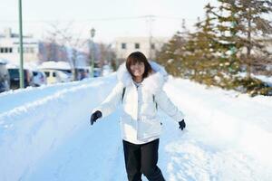 feliz viajante com suéter e mochila caminhando em neve coberto estrada dentro gelado clima, mulher turista passeios turísticos dentro sapporo cidade, Hokkaido, Japão inverno viagem e período de férias conceito foto