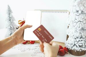 □ Gentil mãos berço uma Smartphone com uma branco tela zombar e dedos em a tela para fazer compras conectados acima de a festivo cena do uma lindo Natal árvore adornado, com Natal bolas, e pinho cones. foto