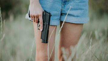 menina dentro jeans calção e com uma arma de fogo dentro dele mão posando dentro a campo. fechar acima foto