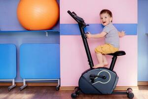 uma pequeno bebê senta em a exercício bicicleta dentro a Academia foto