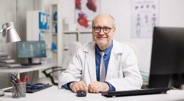 maduro médico vestindo Óculos e sorridente dentro hospital gabinete sentado às escrivaninha. foto