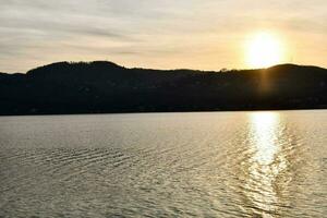 a Sol é configuração sobre uma lago com montanhas dentro a fundo foto