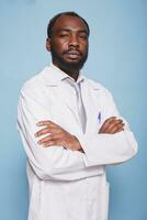 africano americano masculino médico em pé contra a isolado azul fundo. sério Preto homem vestindo uma branco laboratório casaco posando com dele braços cruzado dentro frente do a Câmera. foto