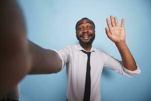 em azul fundo, alegre vlogger ondas Olá para a Câmera durante Video chamada conferência. ponto do Visão do africano americano influenciador levando uma selfie enquanto fazer mão gesto para social meios de comunicação. foto