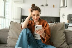 retrato do jovem moderno mulher lendo em Móvel telefone, rolagem social meios de comunicação aplicativo em Smartphone, sentado em sofá dentro vivo quarto dentro casual roupas foto