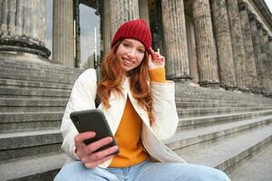 retrato do jovem ruiva mulher dentro vermelho chapéu, sentado em escadaria, turista parece às dela Móvel telefone, descansos em Escadaria do museu, conecta público Wi-fi foto