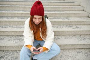 hipster gengibre garota, ruiva mulher senta em escadas com Smartphone, espera para alguém e mensagens em social meios de comunicação em Móvel telefone aplicativo foto