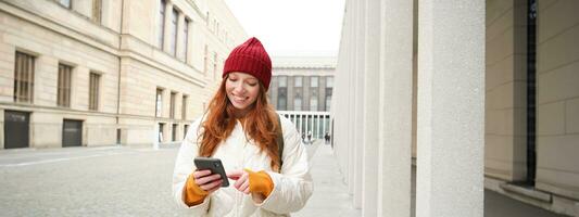 turismo e tecnologia. lindo ruiva menina turista, olhando para uma rota em aplicativo, usando cidade mapa em Smartphone, lendo sobre passeios turísticos lugares em Móvel telefone foto