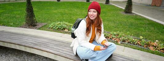 retrato do à moda jovem mulher, 25 anos, senta em Banco dentro parque e usa Móvel telefone, lê conectados notícias, mensagens ou relógios vídeo em Smartphone aplicativo, conecta para público Wi-fi foto