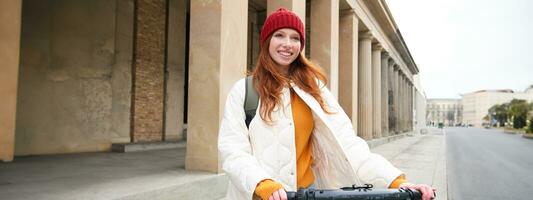 sorridente ruiva europeu menina drives público escooter, turista explora cidade, passeios dentro cidade Centro foto