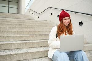 sorridente ruiva garota, aluna senta em escadas ao ar livre e usa computador portátil, conecta para público Wi-fi dentro cidade e trabalho em projeto, usa Internet em computador foto