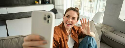 amigáveis sorridente jovem mulher, sentado em sofá e acenando mão às Smartphone Câmera, vídeo bate-papos, falando em Móvel telefone, conecta para conectados conversação em inscrição foto