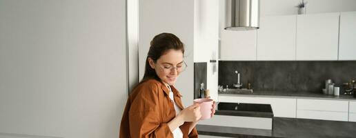 retrato do aconchegante, lindo e relaxado jovem mulher às lar, em pé e inclinado em parede com copo do café, bebendo chá com pensativo face expressão foto