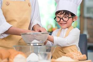 menino bonito asiático e linda mãe peneirar a farinha de massa com peneira peneira na cozinha em casa na mesa para preparar para assar a padaria e o bolo. crianças tailandesas brincando com farinha como chef engraçado foto