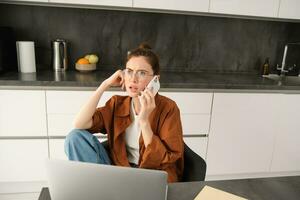 retrato do mulher trabalhando a partir de casa dentro copos, sentado com confuso face depois de respondendo uma telefone chamar, tem computador portátil em mesa foto