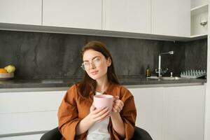 retrato do jovem mulher desfrutando copo do café dentro paz, sentado às lar, segurando caneca com ervas chá, vestindo copos, relaxante às dentro cozinha foto