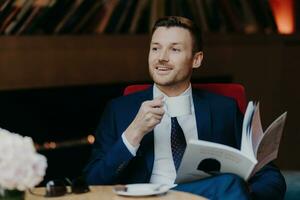 homem dentro terno goza café enquanto leitura, relaxado dentro vermelho cadeira, elegante foto