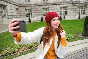 jovem adolescente ruiva menina senta em Banco dentro parque e leva selfie, faz uma foto do ela mesma com Smartphone aplicativo, registros vlog