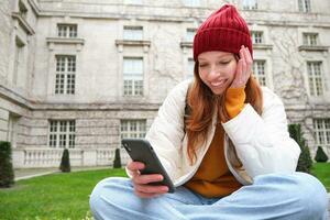 retrato do à moda jovem mulher, 25 anos, senta em Banco dentro parque e usa Móvel telefone, lê conectados notícias, mensagens ou relógios vídeo em Smartphone aplicativo, conecta para público Wi-fi foto