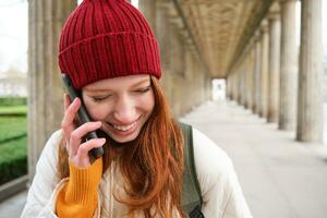 retrato do ruiva europeu menina dentro vermelho chapéu, faz uma telefone chamar, anda em dentro cidade e fala para amigo em Smartphone foto