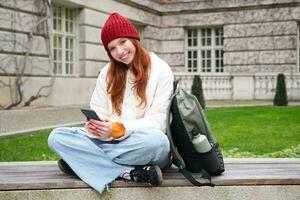 ruiva garota, fêmea aluna senta com Móvel telefone em Banco dentro parj, inclina-se em dela mochila. mulher navegando social meios de comunicação aplicativo alimentação em dela Smartphone foto