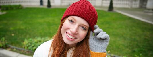 fechar acima retrato do lindo ruiva mulher dentro vermelho tricotado chapéu, caloroso luvas, sorridente e olhando feliz às Câmera, sentado dentro parque foto