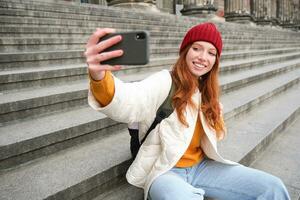 jovem ruiva turista leva selfie dentro frente do museu em escadaria, detém Smartphone e parece às Móvel Câmera, faz foto do ela mesma com telefone