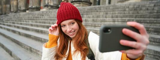 à moda jovem menina dentro vermelho chapéu, leva fotos em Smartphone Câmera, faz selfie Como ela senta em escadas perto museu, posando para foto com aplicativo filtro