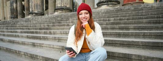 retrato do jovem urbano menina dentro vermelho chapéu, senta em escadas perto museu, detém Móvel telefone, conecta para público Wi-fi e surfa líquido, usa Smartphone apps foto