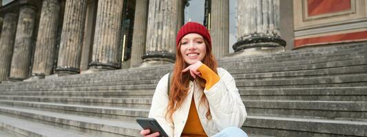 retrato do jovem urbano menina dentro vermelho chapéu, senta em escadas perto museu, detém Móvel telefone, conecta para público Wi-fi e surfa líquido, usa Smartphone apps foto