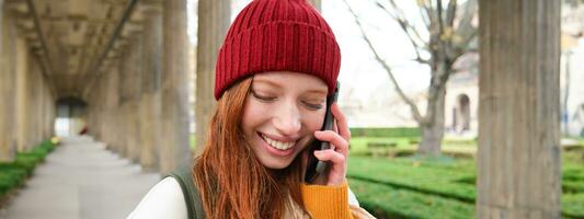 retrato do ruiva europeu menina dentro vermelho chapéu, faz uma telefone chamar, anda em dentro cidade e fala para amigo em Smartphone foto