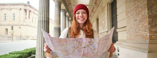 aventureiro ruiva menina anda em dentro Cidade com papel mapa, explora cidade Como turista, parece para popular turismo atrações, parece por aí animado e sorrisos foto