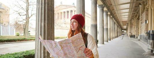 sorridente jovem ruiva mulher dentro vermelho chapéu, parece às papel mapa para Veja para turista atração. turismo e pessoas conceito. menina explora cidade, tentou para encontrar caminho foto