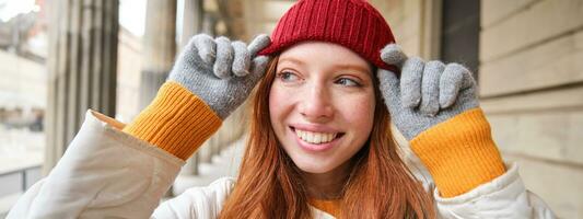 retrato do jovem ruiva mulher dentro tricotado chapéu e luvas, sorrisos e parece aparte, anda em por aí cidade dentro inverno foto