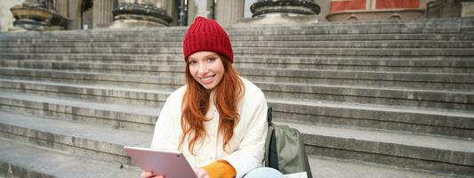 lindo jovem moderno menina com vermelho cabelo, detém digital tábua, senta em escadas perto museu e conecta público Internet, envia mensagem em gadget aplicativo foto