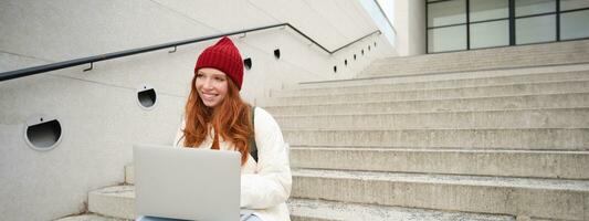 sorridente ruiva garota, aluna senta em escadas ao ar livre e usa computador portátil, conecta para público Wi-fi dentro cidade e trabalho em projeto, usa Internet em computador foto