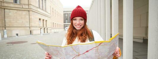 lindo ruiva mulher, turista com cidade mapa, explora passeios turísticos histórico marco, caminhando por aí velho cidade, sorridente alegremente foto