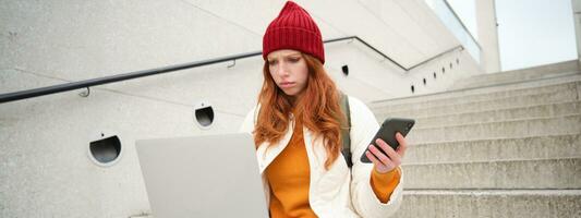 retrato do ruiva mulher senta em escadaria, usa computador portátil e detém Smartphone, parece confuso e chateado às computador tela, tentativas para conectar para Wi-fi público Internet foto