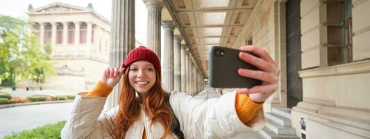 fofa jovem ruiva mulher leva selfie em rua com Móvel telefone, faz uma foto do ela mesma com Smartphone aplicativo em rua