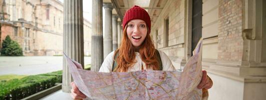 aventureiro ruiva menina anda em dentro Cidade com papel mapa, explora cidade Como turista, parece para popular turismo atrações, parece por aí animado e sorrisos foto