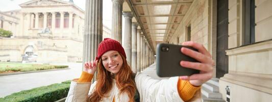 à moda ruiva menina turista, leva selfie dentro frente do turismo atração, faz foto com Smartphone, parece às Móvel Câmera e poses