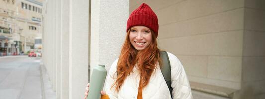 jovem feliz ruiva mulher dentro vermelho chapéu, bebendo a partir de garrafa térmica, aquecimento acima com quente beber dentro dela frasco enquanto caminhando por aí cidade, turista relaxa com caloroso atualizar foto