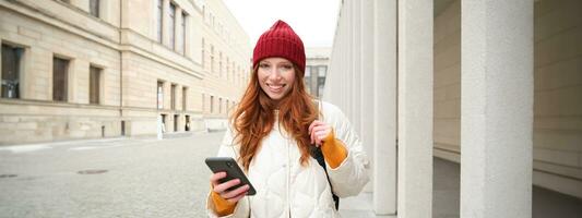 turismo e tecnologia. lindo ruiva menina turista, olhando para uma rota em aplicativo, usando cidade mapa em Smartphone, lendo sobre passeios turísticos lugares em Móvel telefone foto