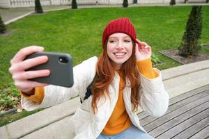 lindo jovem urbano menina leva selfie em Smartphone, relaxado em Banco dentro parque e faz uma foto do ela mesma com Móvel telefone Câmera