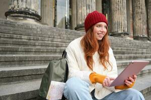 lindo jovem moderno menina com vermelho cabelo, detém digital tábua, senta em escadas perto museu e conecta público Internet, envia mensagem em gadget aplicativo foto