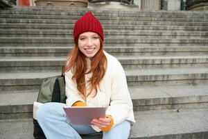 lindo jovem moderno menina com vermelho cabelo, detém digital tábua, senta em escadas perto museu e conecta público Internet, envia mensagem em gadget aplicativo foto